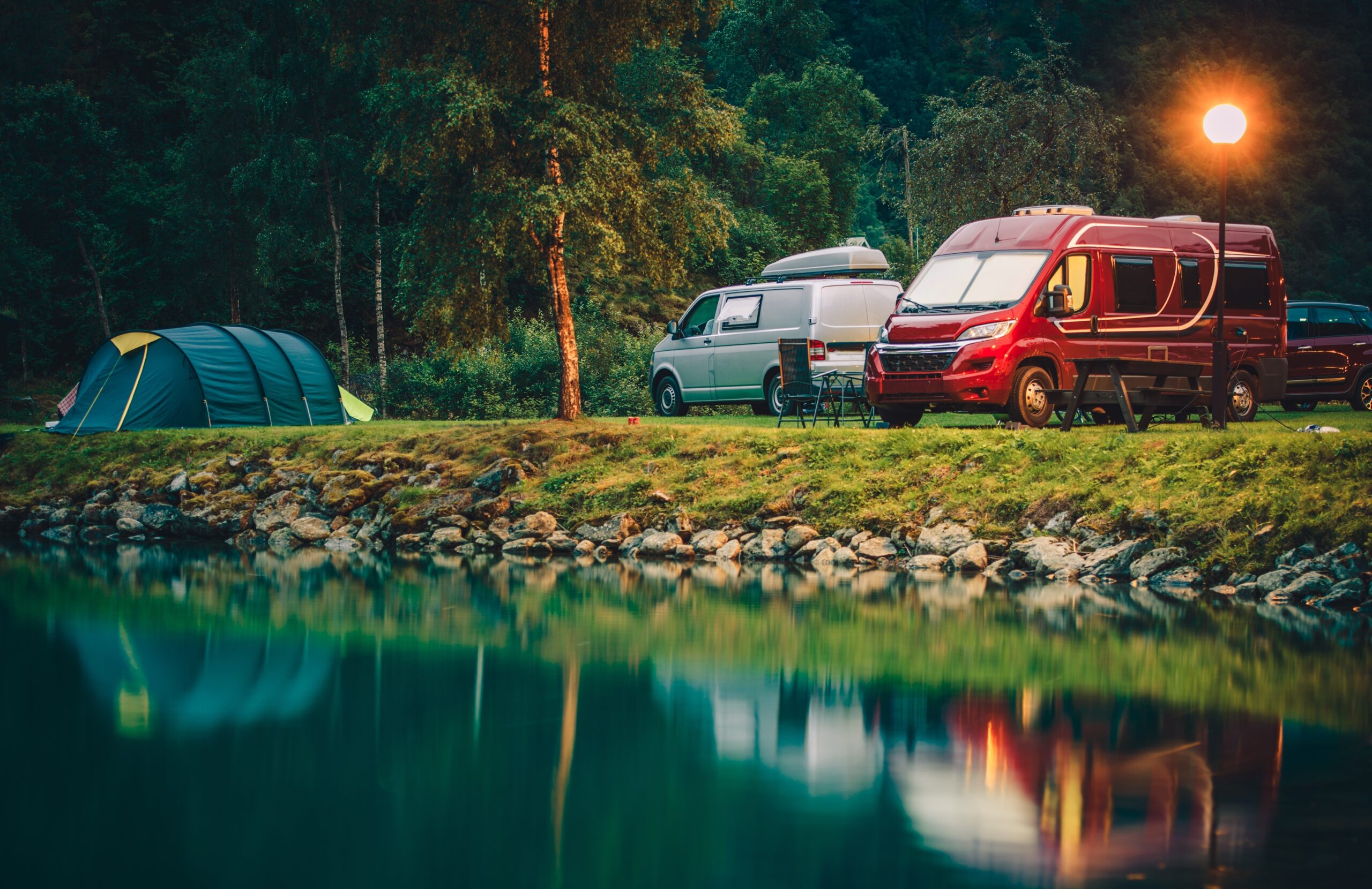 Campsite by a lake with two camper vans, a green tent, outdoor chairs, and a table. Trees and rocks line the shore, and a glowing lamp post lights the scene at dusk. The water reflects the vehicles and surroundings.