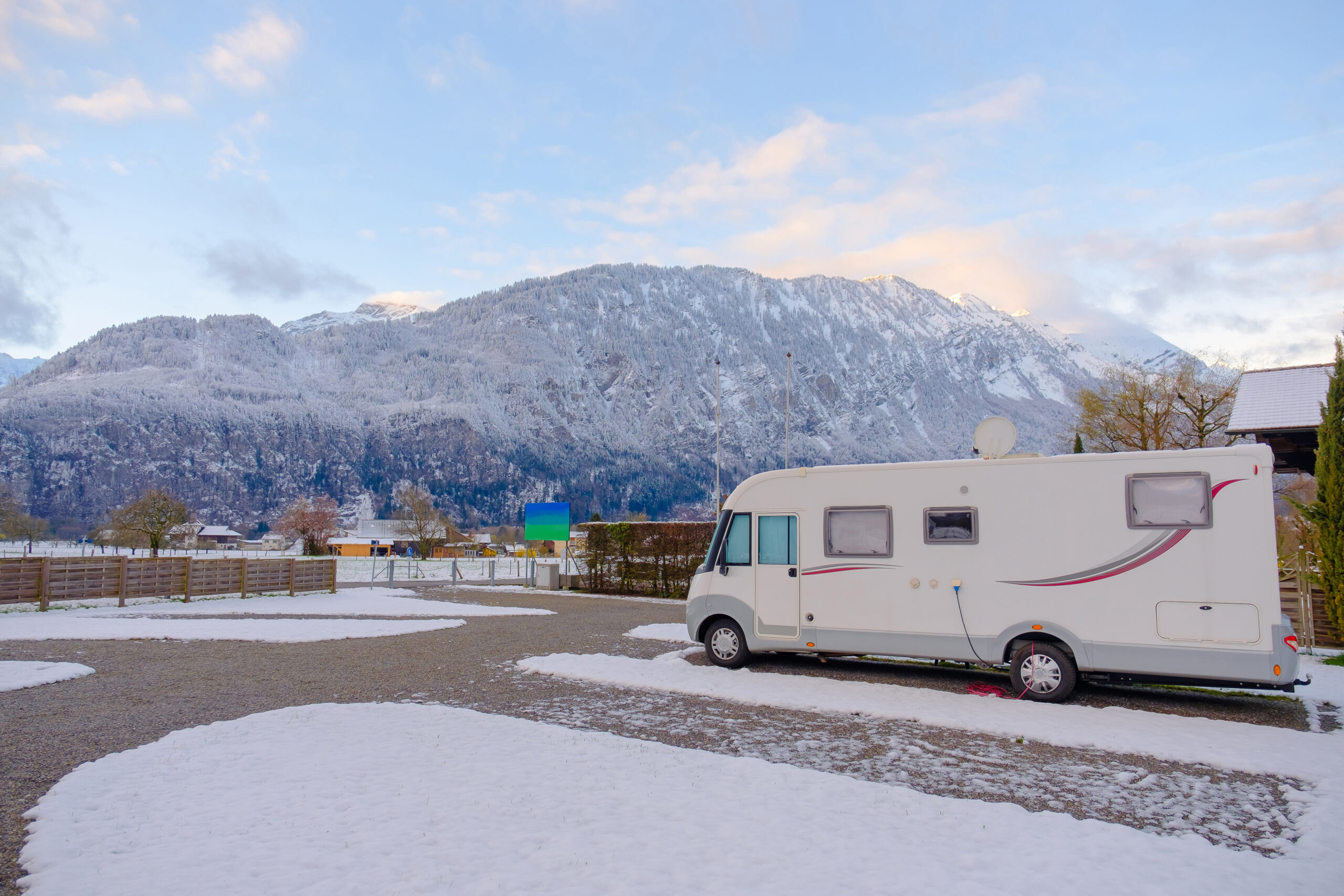 A white camper van is parked on a snowy lot with patches of gravel, with snow-covered mountains and a partly cloudy sky in the background.