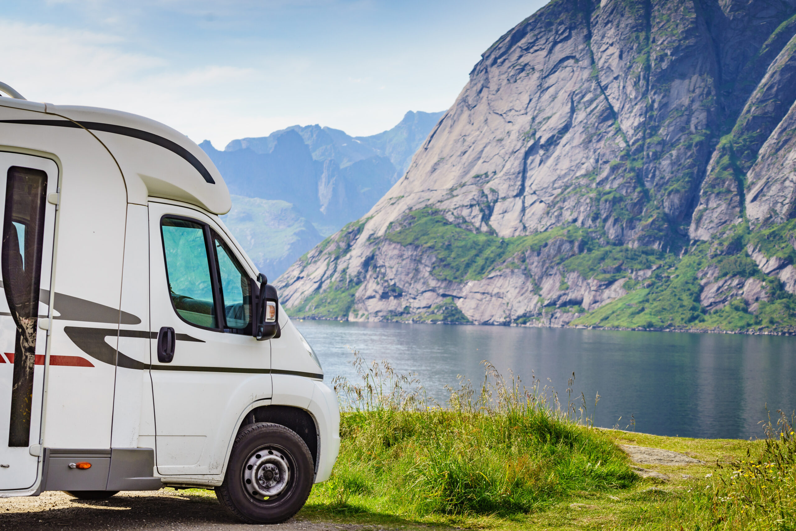 A white camper van parked beside a lake with tall, rocky mountains in the background under a partly cloudy sky, surrounded by green grass and wildflowers.