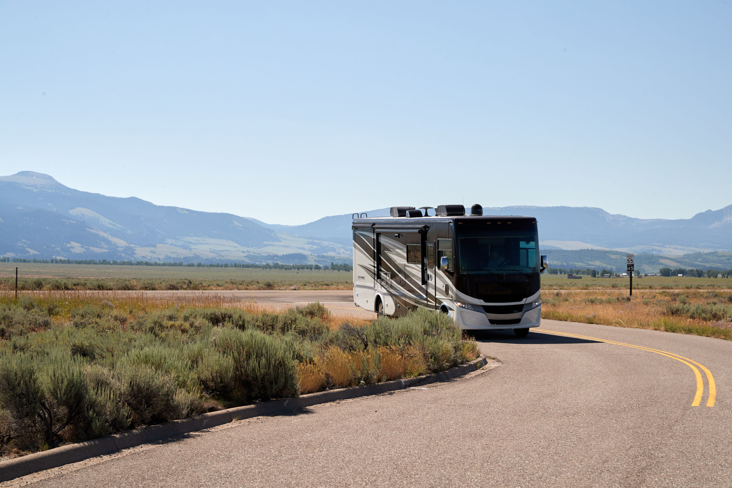 A large motorhome drives on a winding road through a scenic, open landscape with grassy fields and distant mountains under a clear blue sky.